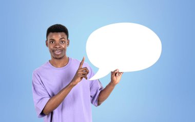 Günlük kıyafet giyen Afrikalı Amerikalı işadamı arka plandaki boş mavi duvarın yanında konuşma baloncuğu ikonu için parmaklarıyla işaret ediyor. Model kavramı, başarılı iş adamı, mesaj