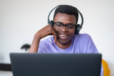 Günlük kıyafet giyen gülümseyen Afrikalı Amerikalı öğrenci, iş yerinde kulaklık ve dizüstü bilgisayarla podcast dinliyor. Uzak, çevrimiçi, uzak öğrenim, iş eğitimi kavramı