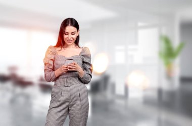 Resmi kıyafet giyen çekici bir iş kadını arka plandaki ofis iş yerinde elinde akıllı telefon tutuyor. Çalışma süreci kavramı, İnternet iletişimi, iş yeri
