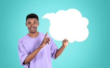 Günlük kıyafet giyen Afrikalı Amerikalı işadamı arka plandaki boş deniz mavisi duvarının yanında konuşma baloncuğu ikonu için parmaklarıyla işaret ediyor. Model kavramı, başarılı iş adamı, mesaj