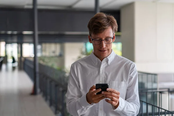 Gözlüklü Genç Avrupalı Işadamı Bulanık Ofis Salonunda Akıllı Telefon Kullanıyor — Stok fotoğraf