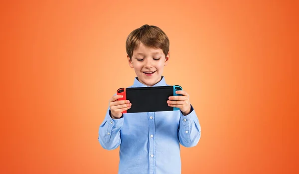 Portrait of cheerful little boy in blue shirt using handheld console over orange background. Concept of gaming and online entertainment