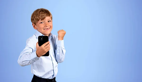 高兴的孩子举着拳头和手机看着相机 复制空旷的蓝色背景 成功和成就的概念 — 图库照片