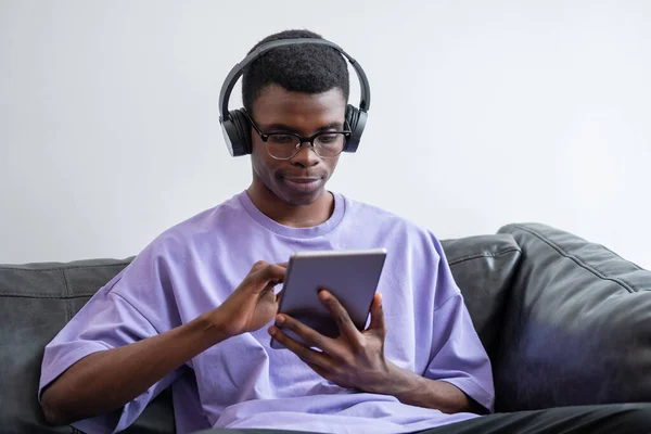 Mladý Afričan Používá Tablet Rukou Sedí Pohovce Poslouchá Hudbu Online — Stock fotografie