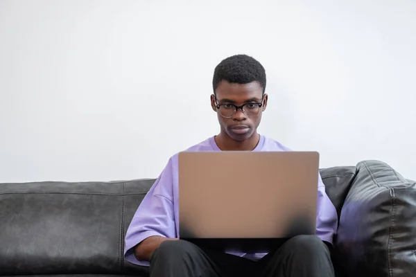 年轻的非洲男子使用笔记本电脑 在沙发上工作 集中的外观和距离的工作 联机服务的概念 — 图库照片