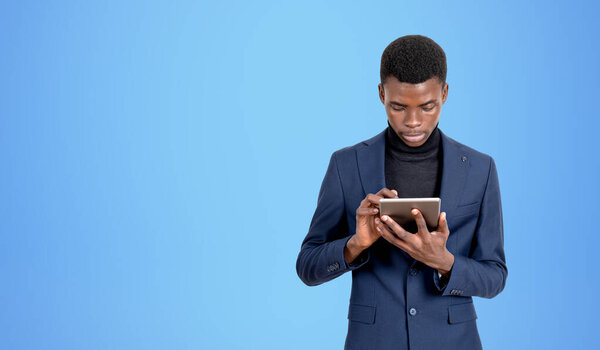 Serious black businessman typing in tablet, concentrated portrait. Online network and social media, copy space empty blue background. Concept of messenger, chat and communication