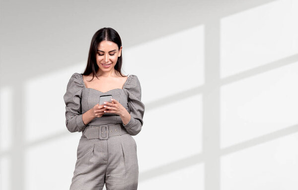 Attractive businesswoman wearing formal wear is standing holding smartphone near empty wall in background. Concept of working process, internet communication, time management