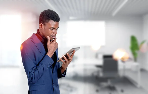 正式なスーツを着たハンサムなアフリカ系アメリカ人ビジネスマンは バックグラウンドでオフィスの職場でタブレットデバイスを見て立っています 現代のガジェットの概念 モバイル通信 時間管理 — ストック写真
