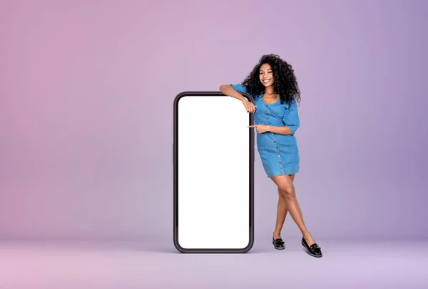 黒の若い女性は フル長さ 紫色の背景に大きなスマートフォンのモックアップを空白の画面を指して笑顔 ソーシャルメディアとコミュニケーションの概念 — ストック写真