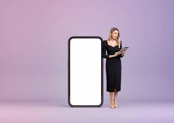 Smiling businesswoman with tablet in hands, full length near large smartphone with mock up copy space display, gradient purple background. Concept of social media