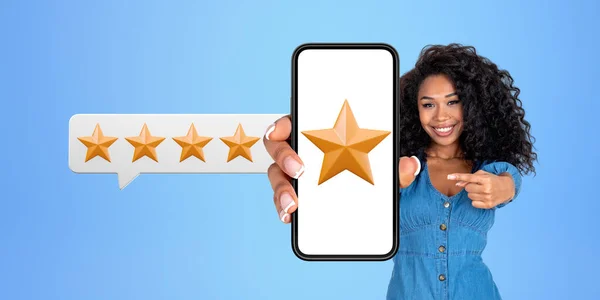 Cheerful young African American woman in casual clothes showing smartphone with five start rating over blue background. Concept of product and service quality ranking