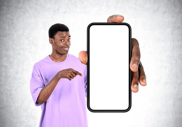 黒の若い男の笑顔 指のコピースペース画面 灰色の具体的な壁の背景をモックアップ大規模なスマートフォンを指しています 接続とソーシャルメディアの概念 — ストック写真