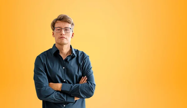 Serious businessman in blue shirt and eyeglasses, looking at the camera, arms crossed on empty copy space orange background. Concept of confidence, leadership and career
