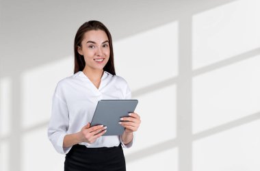Resmi kıyafet giyen gülümseyen çekici iş kadını, arka planda boş beyaz duvarın yanında duran tablet cihazını tutuyor. Model kavramı, başarılı iş adamı, öğrenci, mobil aygıt