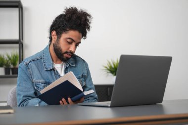 Gündelik kıyafet giyen genç yakışıklı işadamı ofisteki dizüstü bilgisayarın önünde arka planda not alıyor. Çalışma süreci kavramı, uzak çalışma, iş eğitimi