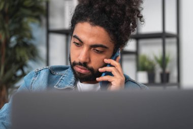 Gündelik kıyafet giyen yakışıklı işadamı arka plandaki ofis bilgisayarının önünde akıllı telefondan konuşuyor. Uzak çalışma kavramı, iş eğitimi, önemli çağrı. Görüntüyü kapat