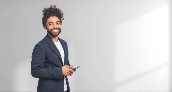 白い壁の近くに立ってスマートフォンを保持エレガントなスーツで陽気な若いアフリカ系アメリカ人ビジネスマンの肖像画 ビジネスコミュニケーションの概念 スペースのコピー — ストック写真