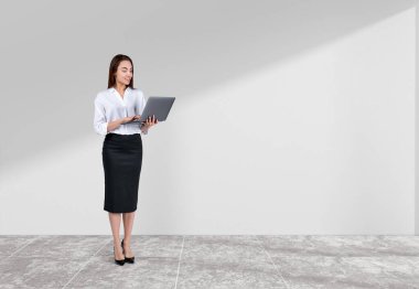 Resmi giysiler içinde gülümseyen çekici iş kadını arka planda beyaz duvarın yanında dizüstü bilgisayarını tutuyor. Beton döşeme. Model kavramı, başarılı iş adamı, çalışan, mobil aygıt