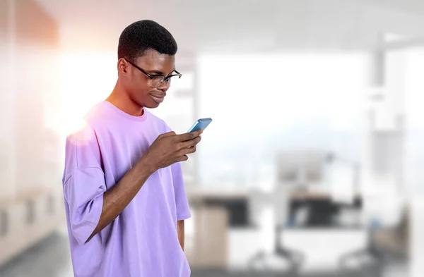カジュアルな服装をしたアフリカ系アメリカ人ビジネスマンがオフィスの職場でスマートフォンを持って立っている 作業プロセスの概念 同僚のためのテキストメッセージ モバイルガジェット — ストック写真
