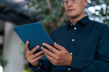 Gözlüklü konsantre işadamı elindeki tablete bakıyor, bulanık ağaçların arka planında alçak açıda duruyor. Uzaklık konferansı, internet ve çevrimiçi ağ kavramı