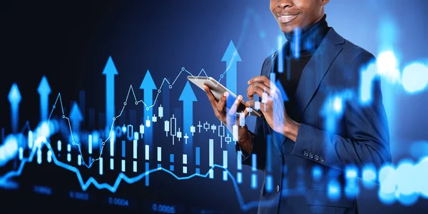 stock image Cheerful young African American businessman using tablet computer over dark blue background with double exposure of growing digital diagrams. Concept of stock market and business success