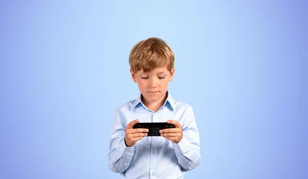 一个严肃的小男孩 穿着正装 拿着智能手机 站在蓝色背景上看屏幕 电子学习和社交媒体的概念 游戏和技术 — 图库照片
