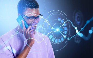 Gülümseyen yakışıklı Afro-Amerikan iş adamı günlük kıyafet giyerek akıllı telefondan konuşuyor. Grafiklerle dijital arayüz, pasta diyagramları, ikili kod, daire hologramı. Başarılı ticaret kavramı