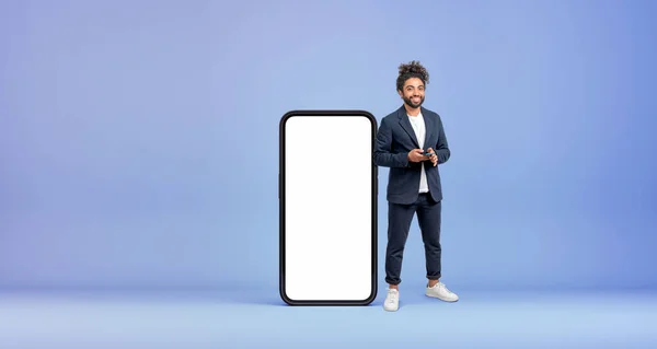 青の背景にモックアップ画面を持つ大きなスマートフォンの近くに立ってスマートフォンを持つ陽気な若い中東のビジネスマン ソーシャルメディアの概念 — ストック写真