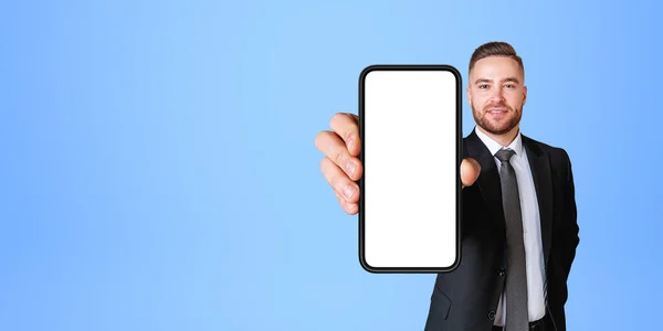 青い背景の上に立って空のモックアップ画面とスマートフォンを示すエレガントなスーツで陽気な若いヨーロッパのビジネスマンの肖像画 コミュニケーションと広告の概念 スペースのコピー — ストック写真