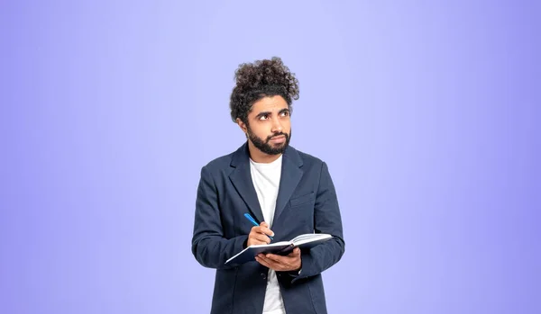 Thoughtful young Middle Eastern man college student standing with notebook and pen and looking sideways and upwards over purple background. Concept of planning, creativity and education