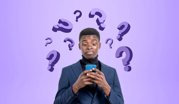 スマートフォンで若い黒人実業家の肖像画は 紫の背景に疑問符のセットと驚きと混乱した アイデアの概念 — ストック写真