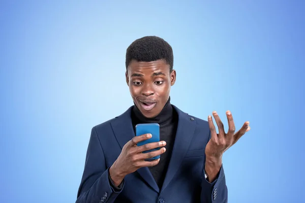 正式なスーツを着たハンサムなアフリカ系アメリカ人ビジネスマンが背景に空の青い壁の近くのスマートフォンで見て立っています 現代のガジェットの概念 モバイル通信 時間管理 — ストック写真