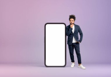 Düşünceli görünümlü, uzun boylu, leylak arkaplanlı akıllı telefon ekranının yanında duran Arap işadamı. Hizmet ve web sitesi kavramı