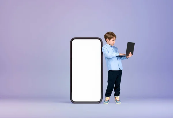 身穿正装的英俊小男孩站在一个大的空白白色模拟智能手机盒旁边拿着笔记本电脑 紫色的墙小玩意的概念 进步的孩子 — 图库照片