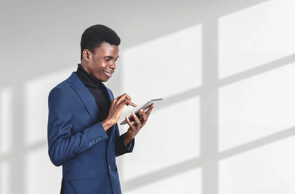 正式なスーツを着たハンサムなアフリカ系アメリカ人実業家が背景に空の白い壁の近くのタブレットデバイスで見て立っている 現代のガジェット モバイル通信 インターネット探検の概念 — ストック写真