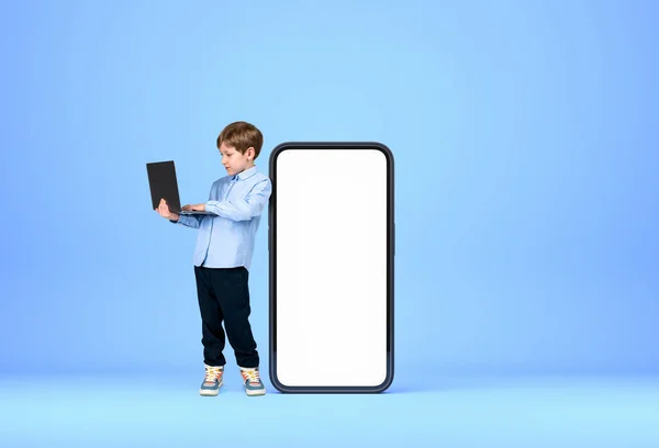 正式な着用を着てハンサムな若い男の子が背景に大きな空の白いモックアップスマートフォンケースの近くにノートパソコンを保持立っている 青い壁だ ガジェット 進歩的な子供の概念 — ストック写真
