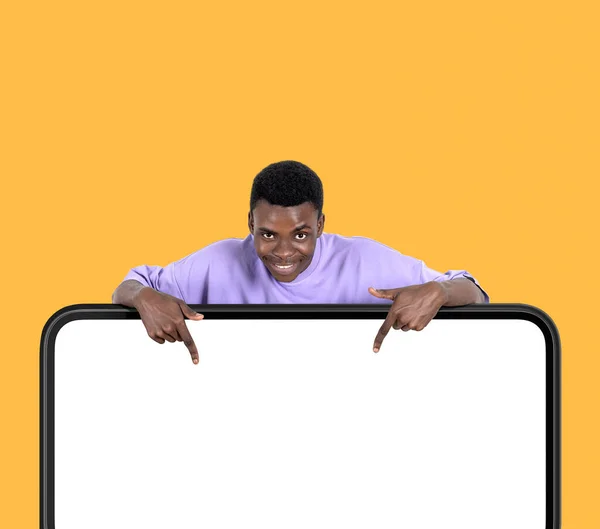 黒人アメリカ人の男が笑顔で 指が黄色い背景に空白のデジタル画面をモックアップで指しています 購読とフォローの概念 — ストック写真