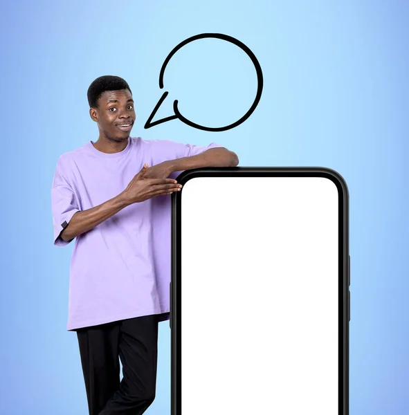 若い黒人男性が笑顔とスピーチバブルコピースペース 青の背景に大きなスマートフォンのモックアップ画面で手を指しています おすすめ おすすめのコンセプト — ストック写真