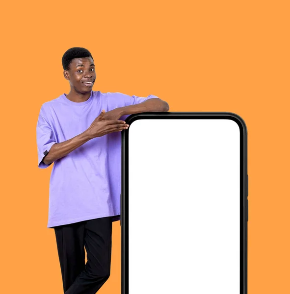 若いアフリカ系アメリカ人の男性は笑顔 手のスマートフォンを指して大規模な空白の画面 オレンジの背景をモック モバイルアプリとオンライン教育の概念 — ストック写真