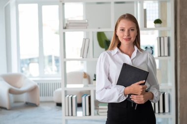 Genç iş kadını öğrenci elinde defterle kameraya bakıp gülümsüyor. Arka planda minimalist ofis odası. İş eğitimi kavramı