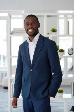 Resmi takım elbiseli gülümseyen Afrikalı Amerikalı işadamı arka plandaki ofis işyerinde el ele poz veriyor. Model, iş adamı, zaman yönetimi, firma, şirket kavramı