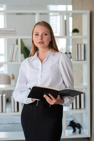 Beautiful businesswoman with opened notebook in hands, concentrated portrait looking at the camera. Minimalist office room on background. Concept of business education