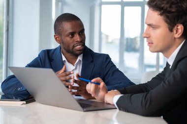 Ciddi bir Afro-Amerikan iş adamı resmi takım elbise giyip iş yerindeki bilgisayardan arka planda belgelerle iş arkadaşlarıyla konuşuyor. Takım çalışması, işbirliği, iş birliği kavramı