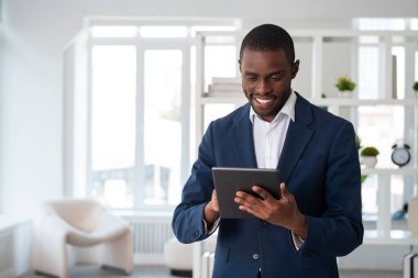 Resmi takım elbise giyen gülümseyen Afro-Amerikan işadamı, ofisindeki tablet cihazında koltuğuyla yazı yazıyor. Model, iş adamı, zaman yönetimi, aygıt, aygıt kavramı