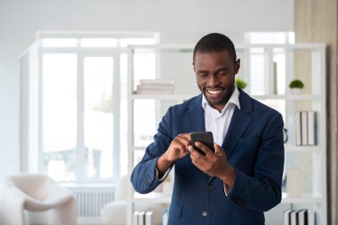 Resmi takım elbise giyen gülümseyen Afro-Amerikan işadamı, ofisinde koltuğuyla akıllı telefonu tutuyor ve daktilo ediyor. Model, iş adamı, zaman yönetimi, aygıt, aygıt kavramı