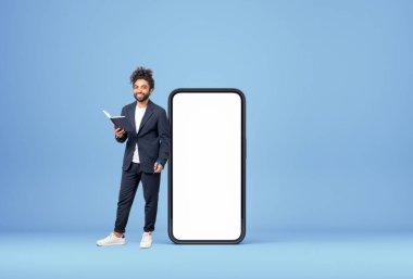 Arap işadamı mutlu görünüş, uzun süre ayakta duran not defteriyle büyük akıllı telefonun yanında boş ekran, mavi arka plan gibi duruyor. E-öğrenim ve ticaret kavramı
