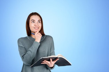 Düşünceli ve güler yüzlü bir kadın elinde defterle tepeden tırnağa bakıp boş mavi arkaplanı kopyalıyor. Düşünce ve fikir kavramı