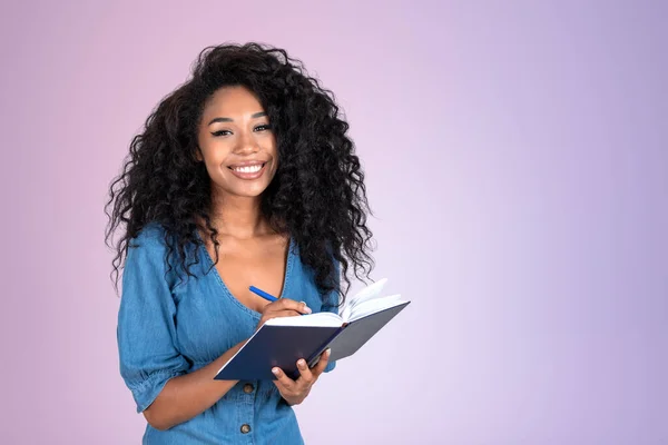 Happy african woman student taking notes, young smiling portrait looking at the camera. Copy space lilac background. Concept of business plan and startup