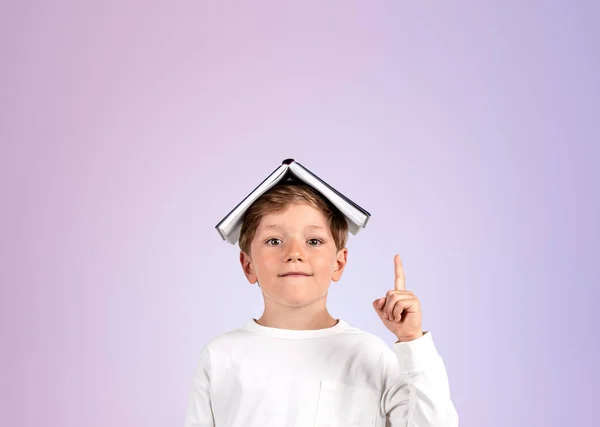 School boy with book on head, index finger pointing up on copy space lilac background. Concept of education and knowledge
