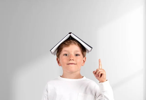 Inspired school boy with a book on head, index finger pointing up on copy space white background. Concept of idea and thoughts
